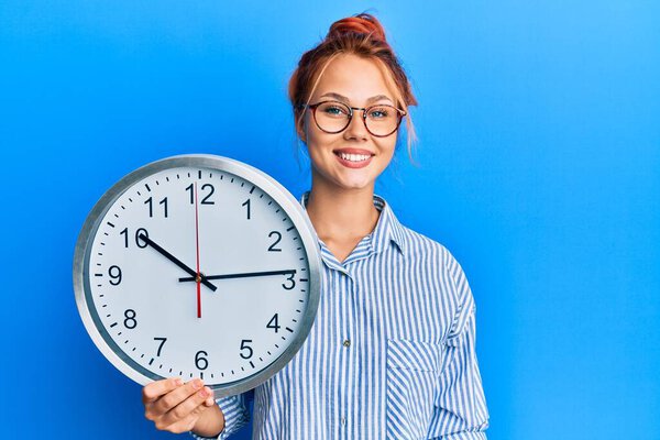 Young redhead woman holding big clock looking positive and happy standing and smiling with a confident smile showing teeth 