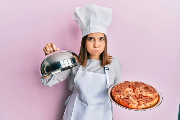 プロの料理人の制服を着た若い白人女性とピザ懐疑的で緊張した帽子を保持し 問題のために動揺して眉をひそめています 否定的な人 — ストック写真