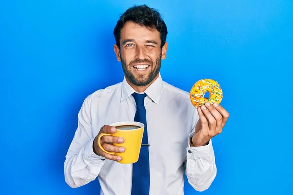 Handsome business man with beard eating doughnut and drinking coffee winking looking at the camera with sexy expression, cheerful and happy face.