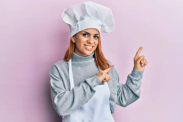 西班牙裔年轻女子身穿职业厨师制服 戴着帽子 面带微笑 看着摄像机 用双手和手指指向旁边 — 图库照片