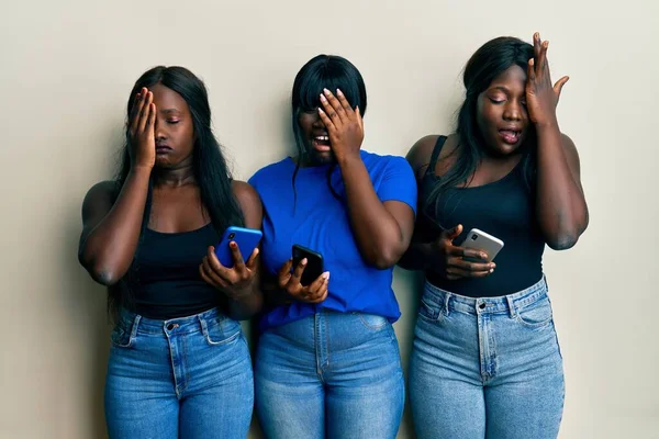 Three young african american friends using smartphone yawning tired covering half face, eye and mouth with hand. face hurts in pain.