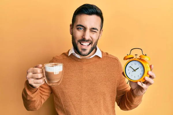 Young hispanic man holding coffee and alarm clock winking looking at the camera with sexy expression, cheerful and happy face. — Stock Image Young Hispanic Man Holding Coffee Alarm Clock Winking Looking Camera Royalty Free Stock Images