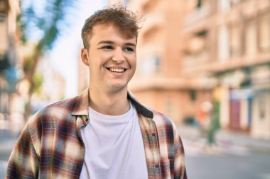 Genç beyaz adam gülümseyerek şehirde mutlu mesut duruyor..