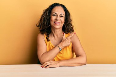 Güzel, orta yaşlı bir kadın, gündelik giysiler giymiş, neşeli bir şekilde masada oturuyor, yüzünde bir gülümseme, mutlu ve doğal bir ifadeyle eli ve parmağı yana doğru işaret ediyor. 