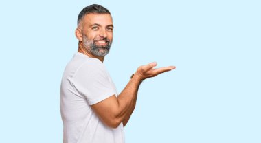 Orta yaşlı beyaz tişört giyen yakışıklı bir adam avuçlarını açıp fotokopi alanını gösteriyor, gülümseyerek mutlu bir gülümseme sergiliyor. 