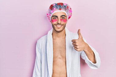 Bornoz ve duş başlığı takan genç İspanyol adam. Göz torbaları kullanıyor. Gülümsüyor, mutlu ve pozitif. 