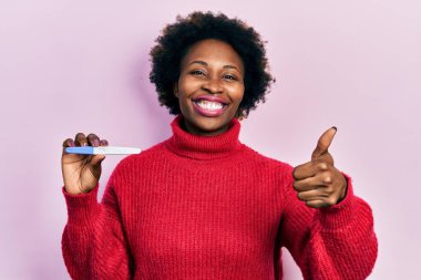 Genç Afrikalı Amerikalı kadın termometreyi tutarken mutlu ve pozitif gülümsüyor, başparmağıyla mükemmel ve onay işareti yapıyor. 