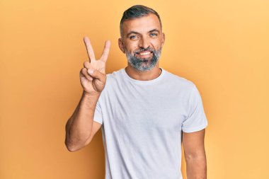 Orta yaşlı, beyaz tişört giyen yakışıklı bir adam iki numarayı gösterip gösterirken kendinden emin ve mutlu bir şekilde gülümsüyor.. 