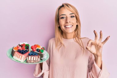 Elinde pasta dilimleri tutan güzel sarışın kadın parmaklarıyla imza atıyor, gülümseyerek mükemmel bir sembol oluşturuyor. 