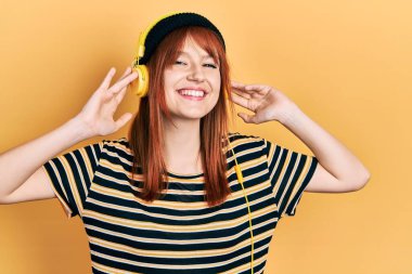 Kızıl saçlı, müzik dinleyen, kulaklık takan mutlu ve havalı bir gülümsemeyle gülümseyen genç bir kadın. diş gösterme. 