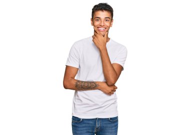 Genç, yakışıklı, Afro-Amerikan bir adam günlük beyaz tişört giyiyor kameraya kendinden emin bakıyor kollarını kavuşturmuş ve çenesini kaldırmış gülümsüyor. Olumlu düşünmek. 