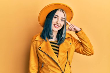 Sarı şapkalı ve deri ceketli genç modern bir kız tek parmakla gülümseyerek, harika bir fikir ya da düşünce, iyi bir hafıza 