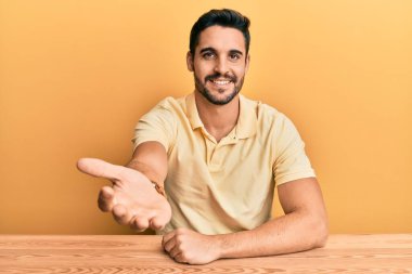 Gündelik giysiler giymiş genç İspanyol bir adam masaya oturup dostça gülümseyerek selamlaşma ve selamlama sunuyor. başarılı iş. 