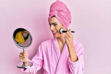 Pembe bornoz giyen genç adam makyaj yapıyor. Yüzü kızarıyor ve kahkahalarla gülüyor.. 