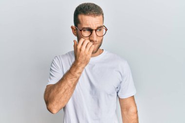 Günlük kıyafetler ve gözlük takan genç beyaz adam gergin ve gergin görünüyor. Ağzı tırnak ısıran elleri var. anksiyete. 