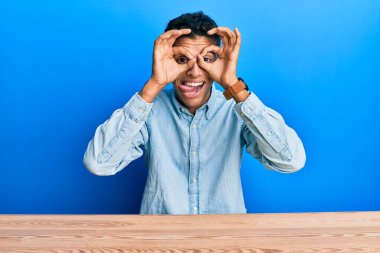Genç, yakışıklı, Afro-Amerikan bir adam, gündelik giysiler içinde masada oturmuş dürbün gibi jest yapıyor, gözleri parmaklarından bakıyor. çılgın ifade. 