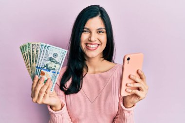 Birleşik Devletler dolarlarını elinde tutan akıllı telefon kullanan genç İspanyol kadın mutlu ve havalı bir gülümsemeyle gülümsüyor. diş gösterme. 