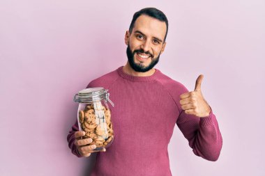 Sakallı genç adam elinde çikolatalı kurabiye kavanozuyla mutlu ve pozitif gülümsüyor, başparmağı mükemmel ve onay işareti yapıyor. 