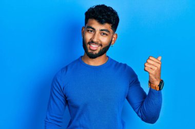 Sakallı, sıradan mavi süveter giyen mutlu yüzlü gülümseyen ve başparmağıyla yüzünü gösteren Arap bir adam.. 