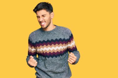 Kış süveteri giyen genç yakışıklı çok mutlu ve heyecanlı kolları havada, gülümseyen ve başarı için bağıran kazanan bir jest yapıyor. kutlama konsepti. 