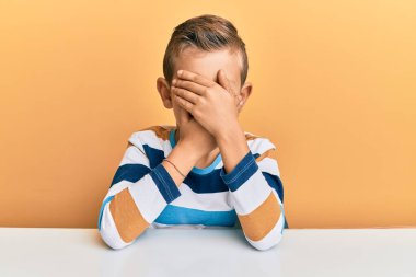 Kafkasyalı sevimli bir çocuk, gündelik giysiler giymiş masaya oturmuş gözlerini ve ağzını elleriyle kapatıyor, şaşırmış ve şok olmuş. duygu gizleme 