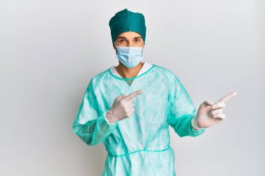 Cerrah üniforması giyen, tıbbi maske takan, gülümseyen ve kameraya bakan genç yakışıklı bir adam.. 