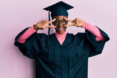 Genç Afrikalı Amerikalı kız mezuniyet şapkası ve tören cübbesi takıyor. Barış sembolü yapıyor. 