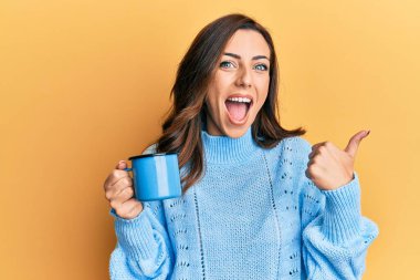 Genç esmer kadın elinde kahve bardağıyla baş parmağını kaldırıyor ve ağzı açık gülümsüyor. 