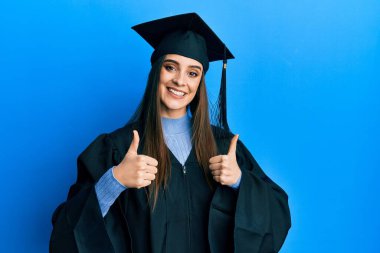 Güzel esmer bir kadın mezuniyet şapkası ve tören cübbesi giyerek başarı işareti yapıyor. Elleriyle olumlu jest yapıyor. Gülümsüyor ve mutlu. Neşeli ifade ve kazanan jest. 