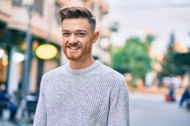 Genç beyaz adam gülümseyerek şehirde mutlu mesut duruyor..