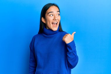 Genç Asyalı kadın, günlük kışlık süveter giyiyor, gülümsüyor, gülümsüyor ve başparmağıyla yan tarafa bakıyor.. 