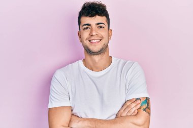 Gündelik beyaz tişört giyen genç İspanyol adam kameraya bakarken kollarını kavuşturup gülümsüyor. pozitif kişi. 