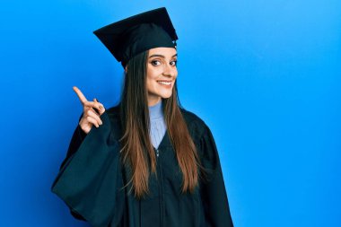 Mezuniyet şapkası ve tören cübbesi takan güzel esmer bir kadın bir numaralı parmağını gösterip onu gösterirken kendinden emin ve mutlu bir şekilde gülümsüyor.. 