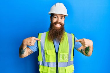 Uzun sakallı, güvenlik kaskı takan, parlak ceketli, kendine güvenen, yüzünde gülümseme olan, kendini gururlu ve mutlu gösteren kızıl saçlı bir adam.. 