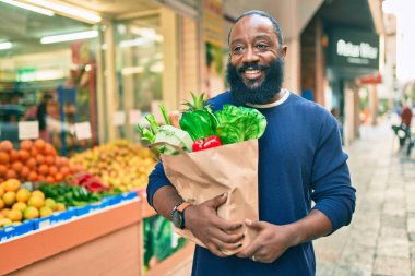 Sakallı Afrikalı Amerikalı bir adam elinde süpermarketten alınmış kağıt poşetler tutuyor.