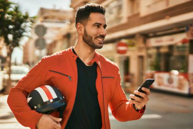 Genç İspanyol adam şehirde moto miğferi tutan akıllı telefon kullanıyor..