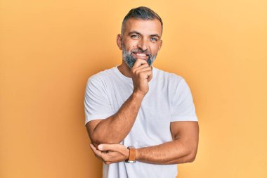 Orta yaşlı beyaz tişört giyen yakışıklı bir adam kameraya kendinden emin bakıyor kollarını kavuşturmuş ve çenesini kaldırmış gülümsüyor. Olumlu düşünmek. 