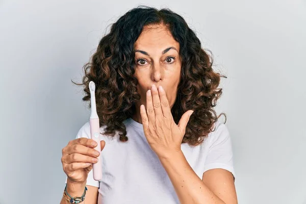 Middle age hispanic woman holding electric toothbrush covering mouth with hand, shocked and afraid for mistake. surprised expression