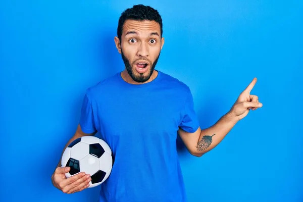 Latynos Brodą Trzymający Piłkę Nożną Zaskoczony Wskazując Palcem Bok Otwarte — Zdjęcie stockowe