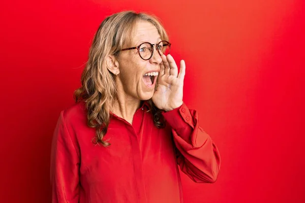 中世のブロンドの女性は 赤い背景の叫びの上に眼鏡をかけ 口の中で手で大声で叫んでいます コミュニケーションの概念 — ストック写真