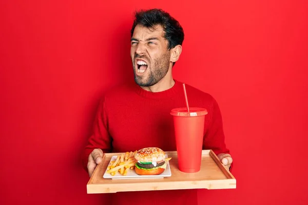 Handsome man with beard eating a tasty classic burger with fries and soda angry and mad screaming frustrated and furious, shouting with anger. rage and aggressive concept.