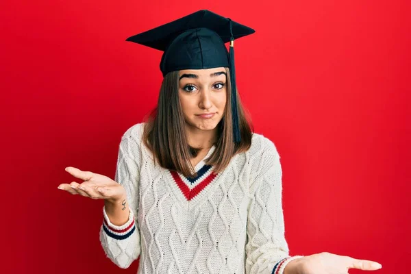 卒業キャップを身に着けている若いブルネットの女の子は 腕や手で混乱した表情を上げました 疑わしい概念 — ストック写真