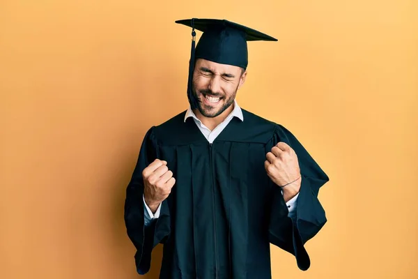 卒業キャップと卒業式のローブを着た若いヒスパニック系の男が腕を上げて成功に興奮し 目は勝利笑顔を祝う閉鎖しました 勝者のコンセプト — ストック写真