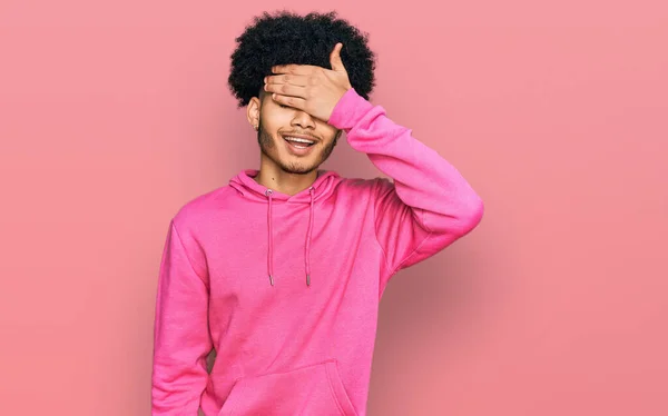 カジュアルなピンクのスウェットシャツを着たアフロヘアの若いアフリカ系アメリカ人男性が笑顔で 驚きのために目をカバー顔に手で笑っています 視覚障害の概念 — ストック写真