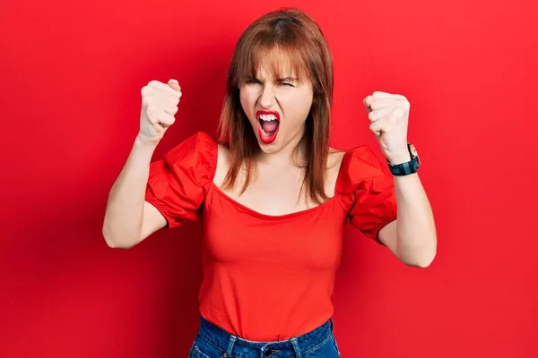 红头发的年轻女人穿着宽松的红色T恤 怒气冲冲地举起拳头 怒气冲冲地大叫着 愤怒和好斗的概念 — 图库照片