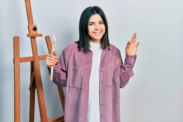 若いブルネットの女性の画家は アートスタジオに座っている指でOkサインを行うアートワードを保持します 笑顔フレンドリーなジェスチャー優れたシンボル — ストック写真