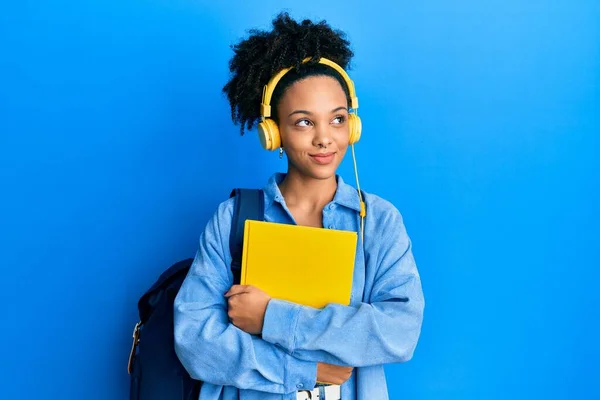 Joven Afroamericana Chica Usando Estudiante Mochila Auriculares Sosteniendo Libro Sonriendo —  Fotos de Stock
