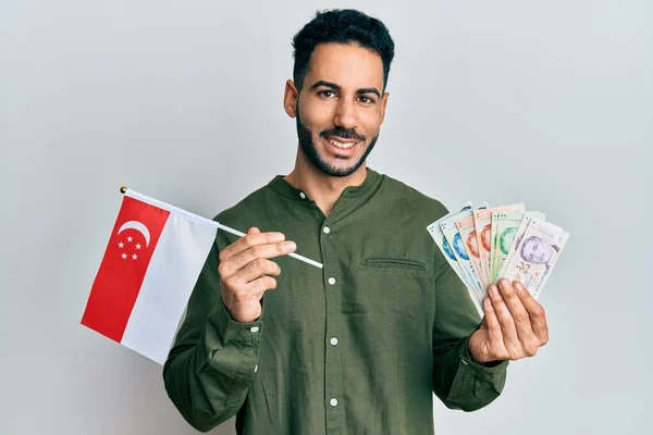 Young hispanic man holding singapore flag and dollars smiling with a happy and cool smile on face. showing teeth.