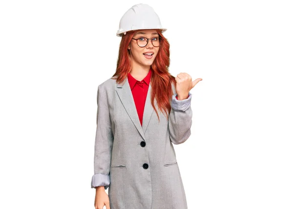 年轻的红头发女人戴着建筑师的硬礼帽 笑容满面 面带微笑 用大拇指指着旁边 — 图库照片