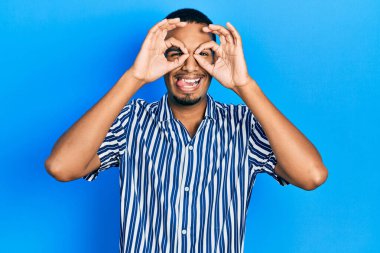 Genç Afro-Amerikan bir adam günlük kıyafetler giyiyor dürbün gibi jest yapıyor, dili dışarı çıkarıyor, gözleri parmaklarından bakıyor. çılgın ifade. 